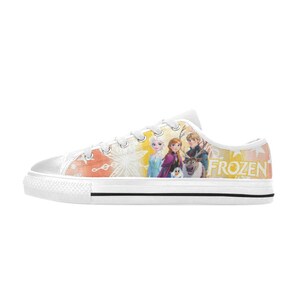 Puede incluir: Zapatillas bajas de lona blanca con un gr&aacute;fico colorido de personajes de la pel&iacute;cula "Frozen". El dise&ntilde;o incluye a Elsa, Anna, Kristoff, Olaf y Sven. La palabra "FROZEN" est&aacute; impresa en amarillo.