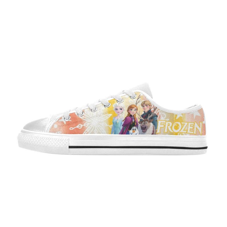 Puede incluir: Zapatillas bajas de lona blanca con un gr&aacute;fico colorido de personajes de la pel&iacute;cula "Frozen". El dise&ntilde;o incluye a Elsa, Anna, Kristoff, Olaf y Sven. La palabra "FROZEN" est&aacute; impresa en amarillo.
