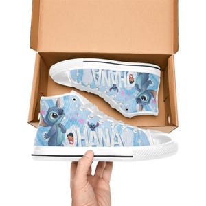 Puede incluir: Zapatillas altas de lona con un dise&ntilde;o azul y blanco con la palabra "OHANA" y un personaje de dibujos animados. Las zapatillas est&aacute;n en una caja de cart&oacute;n, con una zapatilla visible y la otra parcialmente visible.