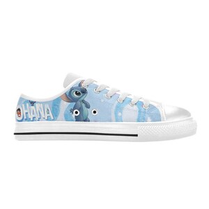 May include: White canvas sneakers featuring a blue and white design with the word "OHANA" and a cartoon character. The shoes have white laces, a white rubber sole, and a black stripe.