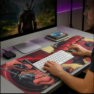 May include: A long rectangular mouse pad featuring a Deadpool graphic in red and black. The pad is on a desk with a white keyboard, mouse, and computer monitor displaying a video game scene. The Deadpool logo is prominently displayed.