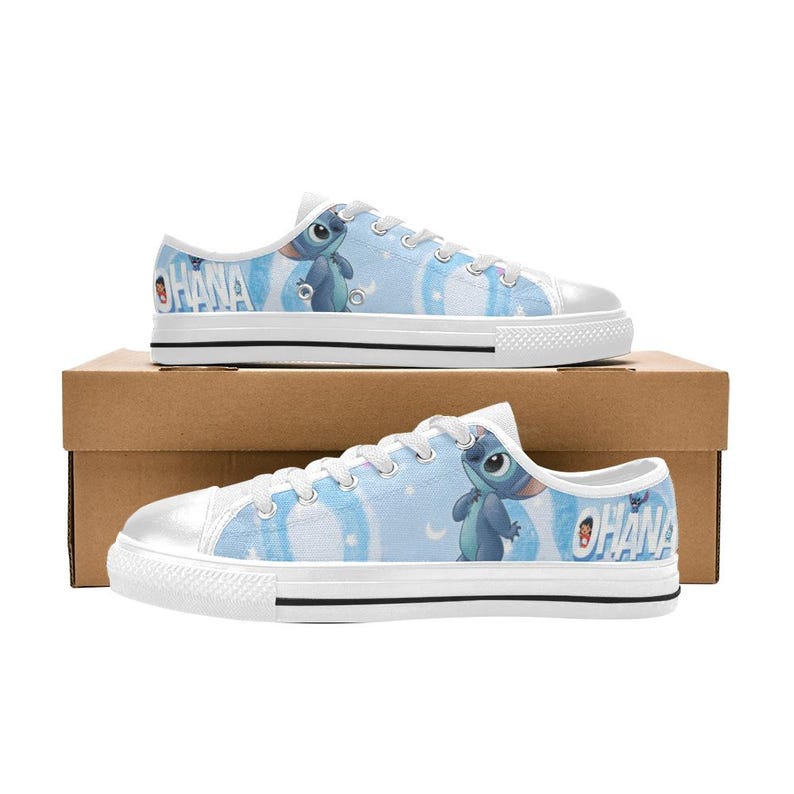 May include: Light blue canvas sneakers featuring a cartoon character and the word "OHANA" in white. The shoes have white laces, a white rubber toe cap, and a white sole. The background of the shoes is a light blue with white clouds.