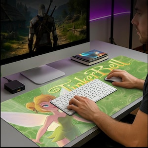 Op de afbeelding: Groene muismat met Tinkerbell en de tekst "Tinker Bell" in goud. De muismat ligt op een bureau met een toetsenbord, muis en computermonitor waarop een videogame wordt weergegeven. De afbeelding toont een persoon die de computer gebruikt.