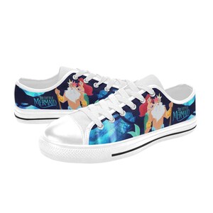 May include: White-laced canvas sneakers featuring a colorful illustration of characters from "The Little Mermaid" on a dark blue background. The design includes Ariel, King Triton, and the movie title. The shoes have a white toe cap and sole.