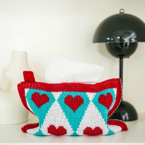 May include: A knitted tissue box cover in red, white, and turquoise, decorated with heart and diamond patterns. The cover is filled with white tissues and sits on a white shelf. A black lamp and white decorative vases are in the background.