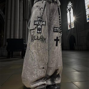Op de afbeelding: Grijze, oversized denim jeans met witte verfspatten en patches met kruisen en tekst. De jeans heeft een zwarte riem met kettingdetails en wordt gedragen met zwarte plateau laarzen. De tekst op de jeans luidt "LEAVE ME ALONE" en "KILL HIM".