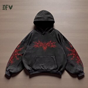 May include: Dark grey hoodie with a hood and front pocket. It has a red, spiky design on the front and sleeves. The upper left corner displays the letters "DFV" with a decorative symbol.