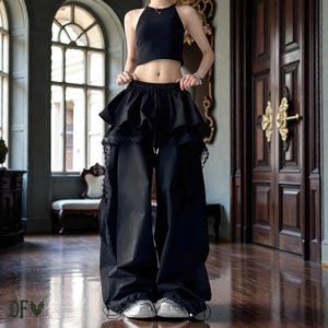 Op de afbeelding: Zwarte haltertop en broek met wijde pijpen en een gelaagd ruchesontwerp. De broek heeft een taille met trekkoord en kanten afwerking. De outfit wordt gecombineerd met zilveren sneakers. De foto is binnenshuis genomen met een houten vloer en grote boogvensters.