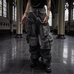 Puede incluir: Pantalones cargo de mezclilla gris oscuro con múltiples bolsillos y un ajuste relajado. Los pantalones tienen una cintura alta y se usan con botas negras. La imagen está tomada en una iglesia con vidrieras.