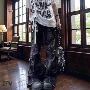 Könnte beinhalten: Schwarze Jeanshose mit Used-Details, Cargotaschen und ausgestelltem Bein. Die Hose hat einen rohen Saum und wird mit Plateauschuhen getragen. Ein weißes T-Shirt mit grafischem Text ist sichtbar.