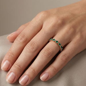 May include: A gold ring with a series of emerald-green gemstones set in a row. The ring is on a person's finger, with the hand resting on a neutral-toned surface. The ring's design is simple and elegant.
