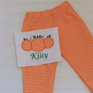 May include: Orange and white checkered pants with a white fabric panel featuring an embroidered design of three pumpkins and the name "Kiley".