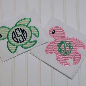 May include: Two embroidered sea turtle designs on white fabric. One turtle is green with a monogram in the center, and the other is pink with a green swirl design.