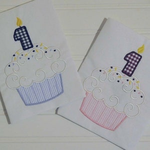 May include: Two white embroidered baby onesies with a cupcake design. One onesie has a blue and white striped cupcake with a purple and white checkered number one candle. The other onesie has a pink and white striped cupcake with a purple and white checkered number one candle.