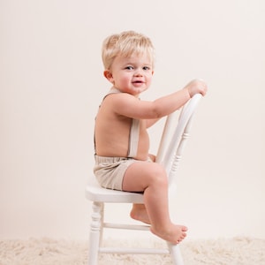 May include: A young boy with blonde hair sits on a white wooden chair. He is wearing a light brown pair of shorts with suspenders. He is looking at the camera and smiling.