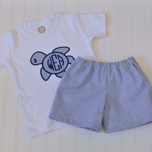 May include: A white short-sleeved t-shirt with a blue embroidered turtle and the monogram "WEA". The shirt is paired with blue and white striped shorts.