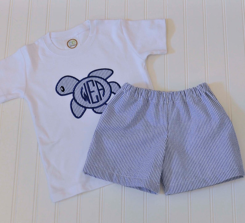 May include: A white short-sleeved t-shirt with a blue embroidered turtle and the monogram "WEA". The shirt is paired with blue and white striped shorts.