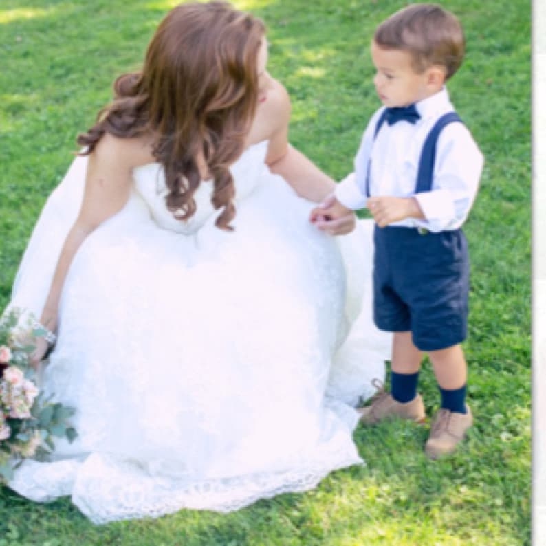 May include: A bride in a white lace wedding dress smiles down at a young boy wearing a white shirt, a navy blue bow tie, and navy blue shorts with suspenders. The boy is looking up at the bride.