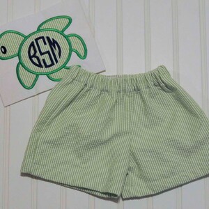 May include: Green and white striped shorts with a white shirt featuring an embroidered green sea turtle with the initials "BSM" in a navy blue circle.