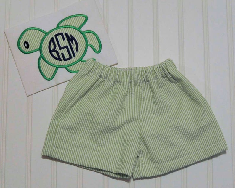 May include: Green and white striped shorts with a white shirt featuring an embroidered green sea turtle with the initials "BSM" in a navy blue circle.