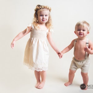 May include: A young girl in a white dress with a flower crown on her head holds hands with a young boy wearing beige shorts and suspenders. They are both smiling and looking at the camera.