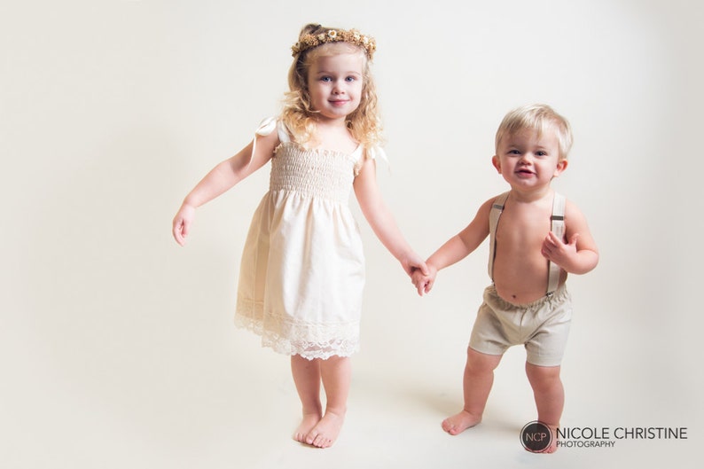 May include: A young girl in a white dress with a flower crown on her head holds hands with a young boy wearing beige shorts and suspenders. They are both smiling and looking at the camera.