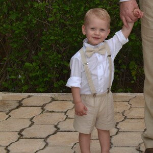 May include: A young boy wearing a white button-down shirt with a beige bow tie and matching shorts. He is wearing beige suspenders and brown shoes. He is holding the hand of an adult.
