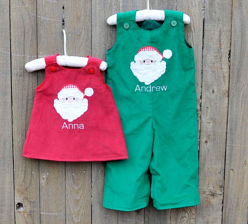 May include: Two personalized Christmas outfits for children. A red dress with a Santa Claus applique and the name "Anna" embroidered on the front. A green overall with a Santa Claus applique and the name "Andrew" embroidered on the front.