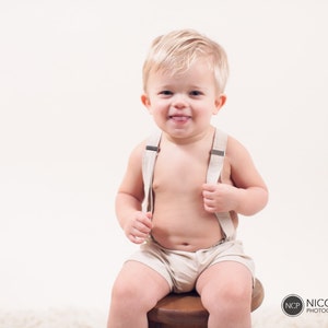 May include: A young child wearing a beige romper with suspenders sits on a wooden stool. The child is smiling and sticking their tongue out.