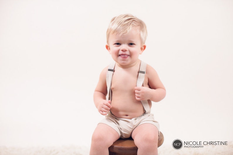 May include: A young child wearing a beige romper with suspenders sits on a wooden stool. The child is smiling and sticking their tongue out.