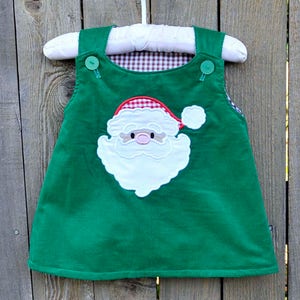 May include: A green corduroy overall dress with a white Santa Claus applique. The Santa has a red hat with white trim and a white beard. The dress has two buttons on the straps.