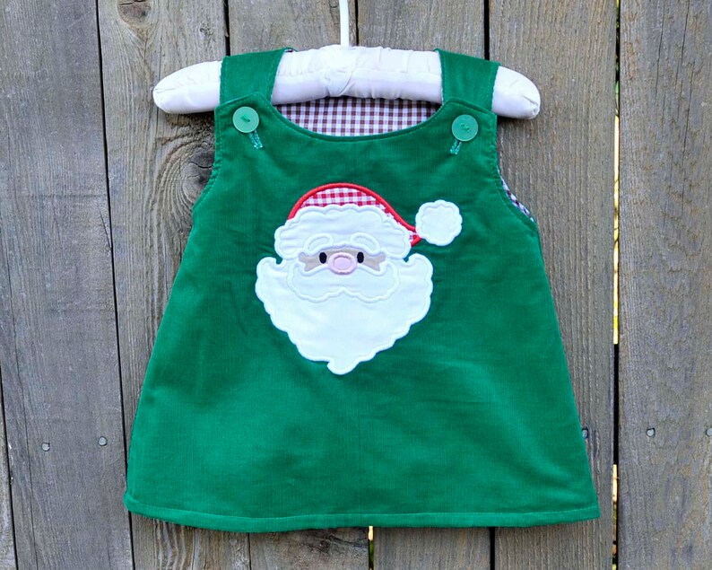 May include: A green corduroy overall dress with a white Santa Claus applique. The Santa has a red hat with white trim and a white beard. The dress has two buttons on the straps.