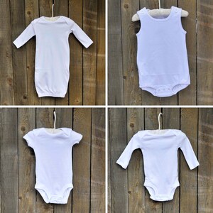 May include: Four white baby onesies hanging on hangers against a wood background. Two are long-sleeved and two are short-sleeved.