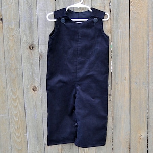 May include: Black corduroy overalls with button closures. The overalls are hanging on a white hanger against a wood background.
