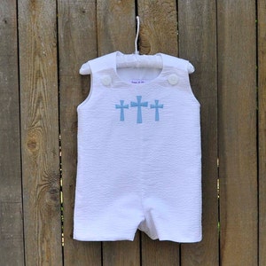 May include: White baby onesie with blue embroidered crosses. The onesie has a button closure at the shoulder and is hanging on a white hanger.