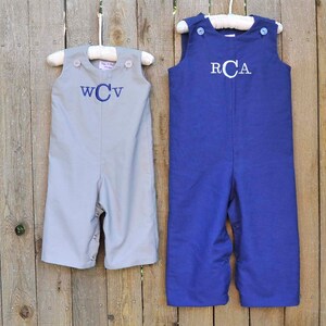 May include: Two pairs of gray and blue overalls with white buttons and embroidered initials. The overalls are hanging on hangers against a wooden fence.