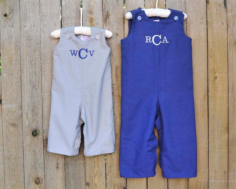 May include: Two pairs of gray and blue overalls with white buttons and embroidered initials. The overalls are hanging on hangers against a wooden fence.