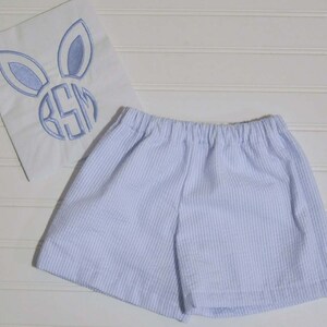 May include: Light blue and white striped shorts with an elasticated waistband. A white square with a blue embroidered bunny face and monogram is next to the shorts.