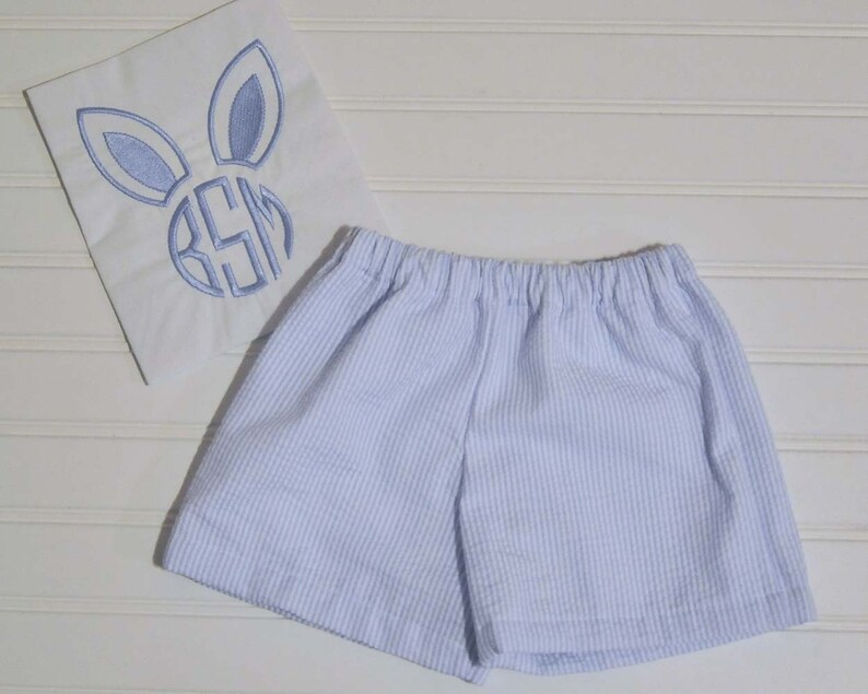 May include: Light blue and white striped shorts with an elasticated waistband. A white square with a blue embroidered bunny face and monogram is next to the shorts.
