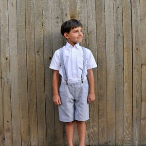 May include: A young boy wearing a white button-down shirt with gray suspenders and matching gray shorts. He is standing in front of a wooden fence.