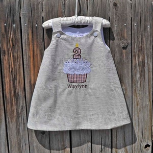 May include: A white and brown gingham dress with a cupcake design embroidered on the front. The cupcake has a white frosting with a yellow candle and a number "2" on top. The dress also has the name "Waylynn" embroidered below the cupcake.