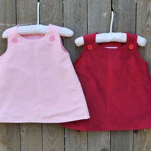 May include: Two sleeveless dresses hanging on white hangers. The dress on the left is pink and the dress on the right is red. Both dresses have two buttons on the front.