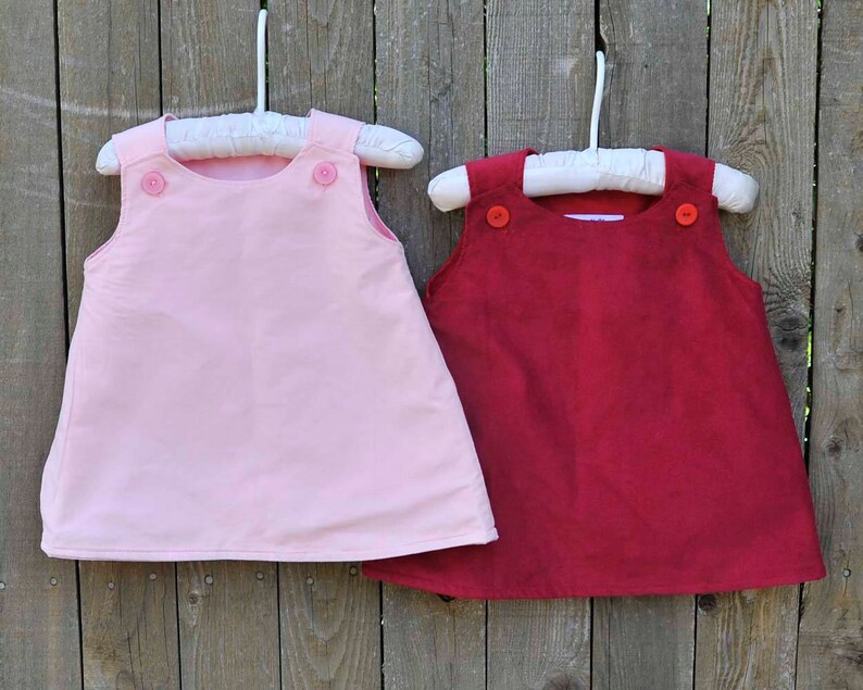 May include: Two sleeveless dresses hanging on white hangers. The dress on the left is pink and the dress on the right is red. Both dresses have two buttons on the front.