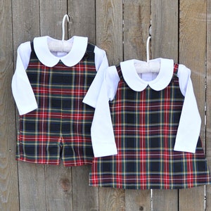 May include: Two plaid outfits for babies, one in a short-sleeved romper style and the other in a long-sleeved dress style. Both outfits feature a white collared shirt with long sleeves and a black, red, and yellow plaid pattern.