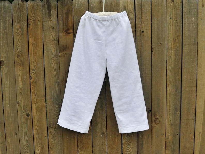 May include: White linen pants hanging on a wooden fence. The pants have a wide leg and a simple design.