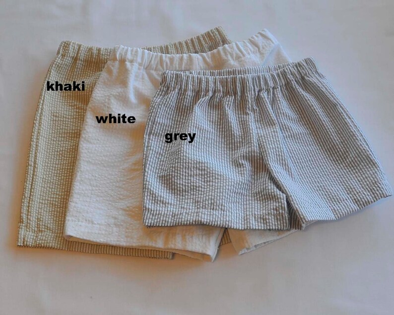 May include: Three pairs of shorts in khaki, white, and grey. The shorts are made of a lightweight fabric with a subtle texture. The shorts have a relaxed fit and are perfect for warm weather.