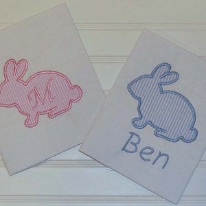 May include: Two white fabric squares featuring embroidered bunny designs. One bunny is pink with a pink letter "M", the other is blue with the name "Ben" embroidered in blue. The bunnies have a striped pattern.