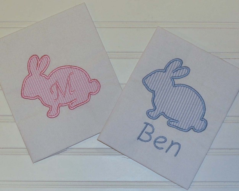 May include: Two white fabric squares featuring embroidered bunny designs. One bunny is pink with a pink letter "M", the other is blue with the name "Ben" embroidered in blue. The bunnies have a striped pattern.