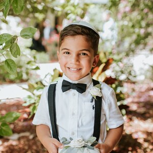 May include: A young boy in a white shirt, black bow tie, and black suspenders holds a white pillow with a white flower and green leaves. He is smiling and wearing a white yarmulke.