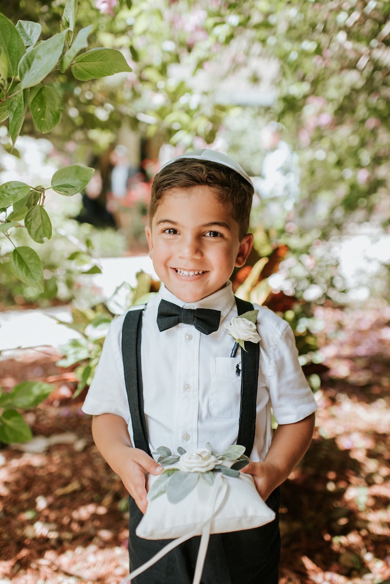 May include: A young boy in a white shirt, black bow tie, and black suspenders holds a white pillow with a white flower and green leaves. He is smiling and wearing a white yarmulke.